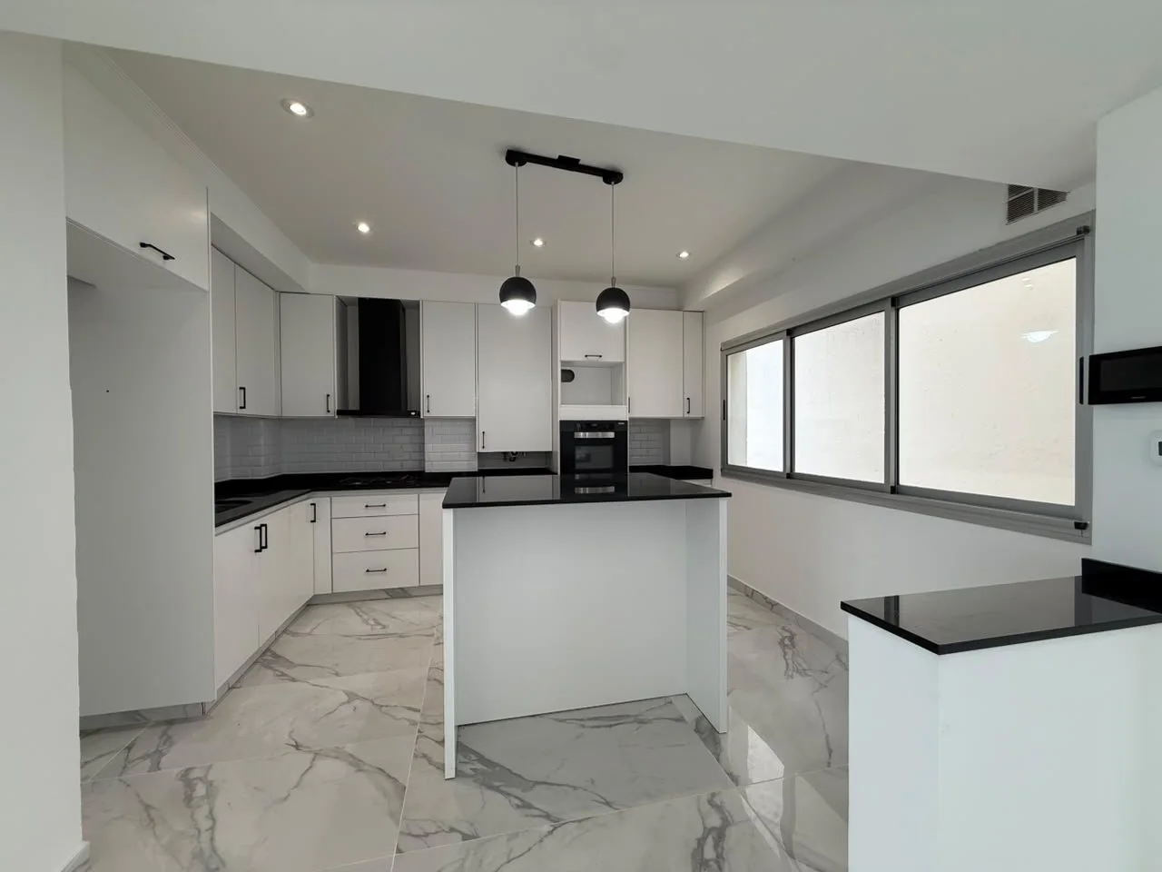 La cocina con isla: mesada de granito, muebles blancos, campana, horno empotrado y ventanal