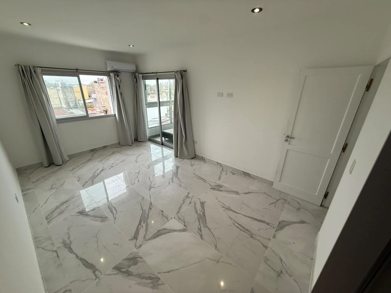 El dormitorio principal desde otro ángulo, con ventana, cortinas, aire acondicionado y puerta corrediza al balcón
