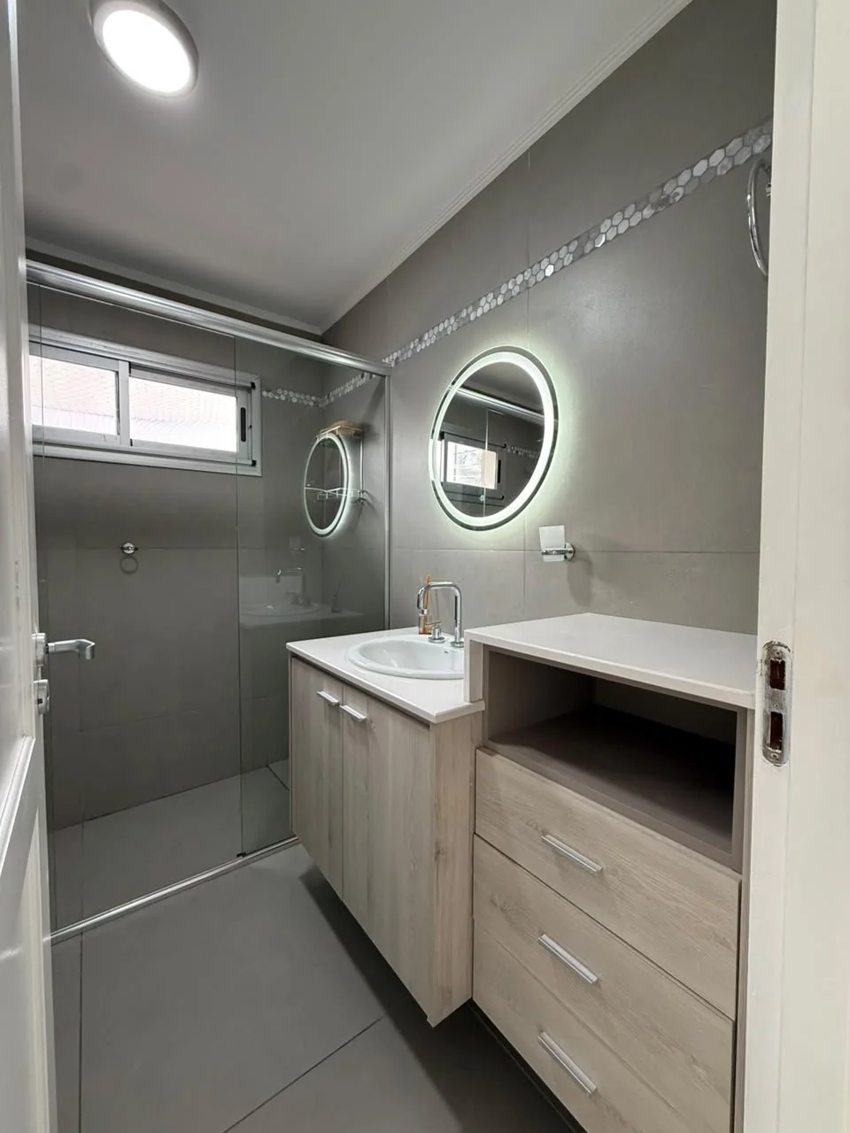 Baño con espejo redondo iluminado, vanitory con cajonera y ducha con mampara