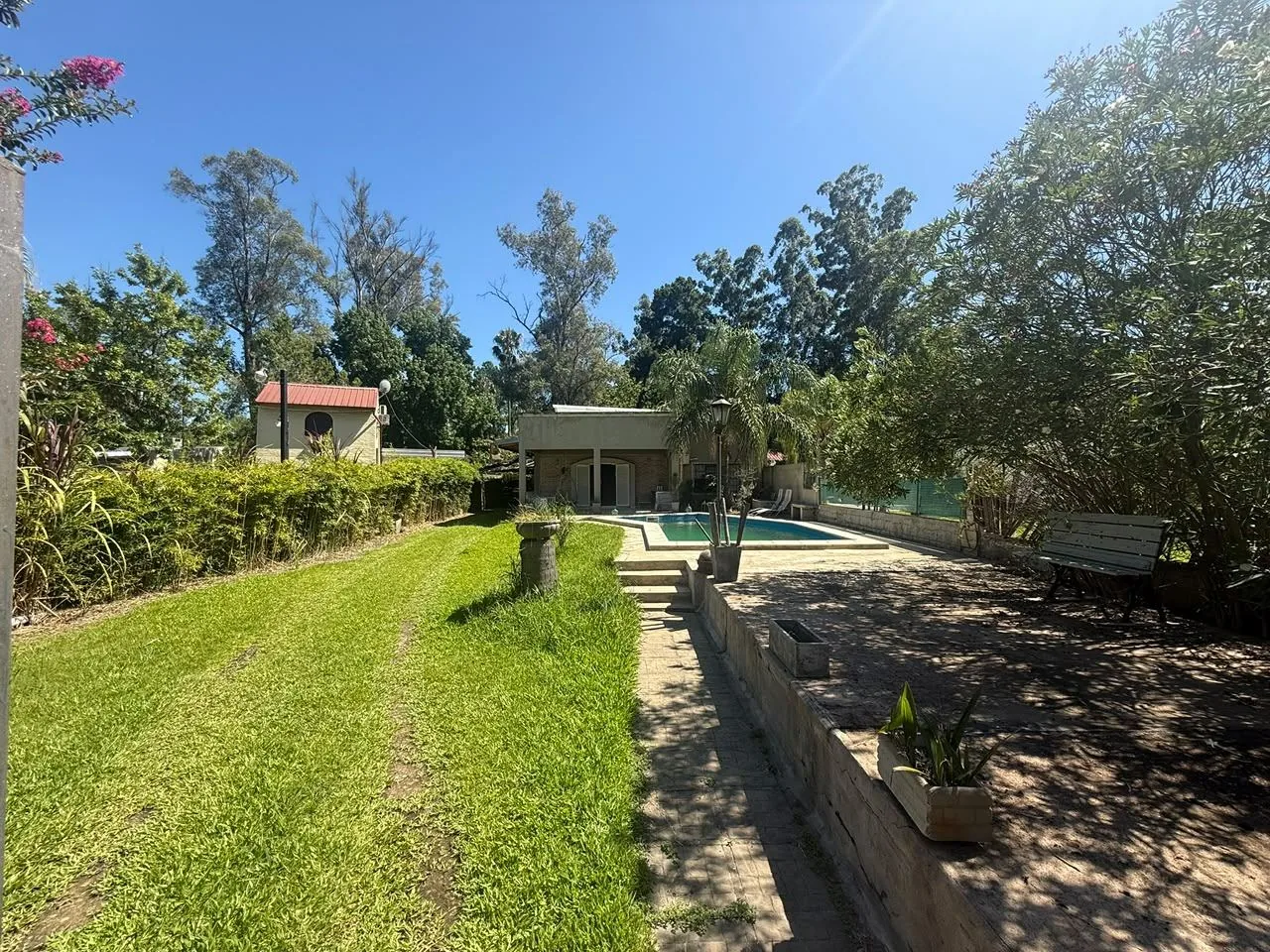 El patio grande: césped, canteros y sombra, con la pileta y la casa en el fondo del terreno