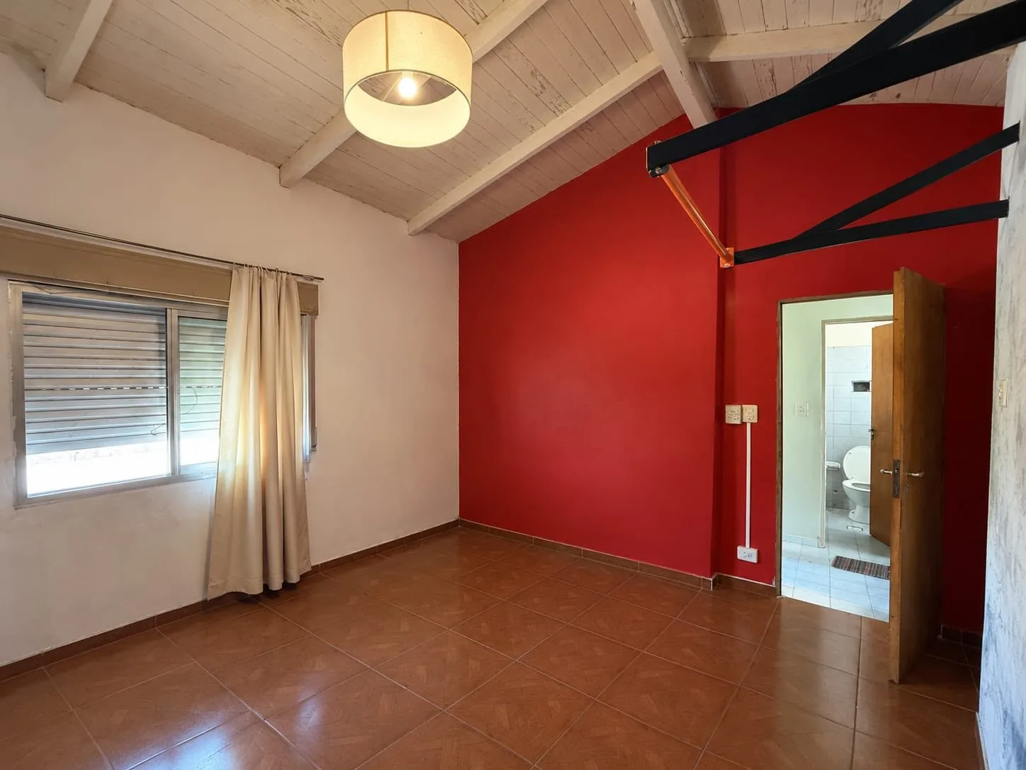 Dormitorio de planta alta, con techo de madera a la vista, ventana con persiana y la puerta hacia el baño